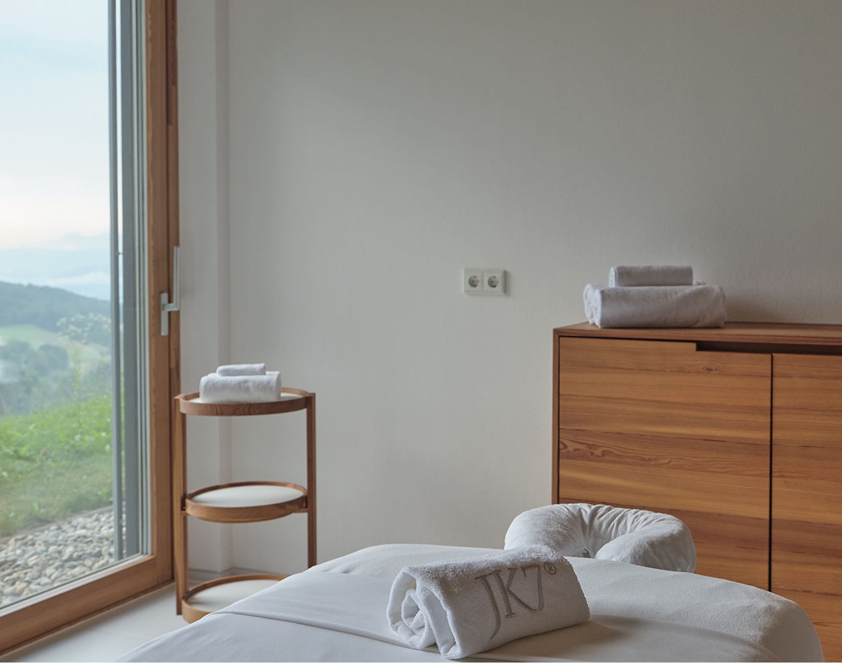 Minimalist treatment room with massage table, folded towels, and a view of nature.